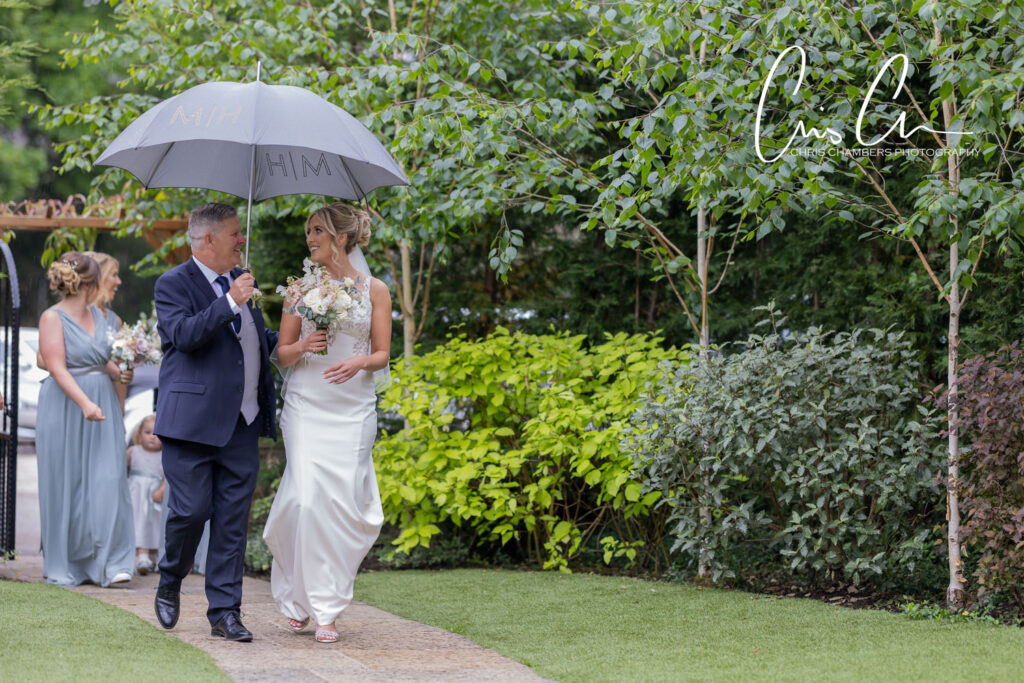 Bride and father walking under umbrella, garden wedding.