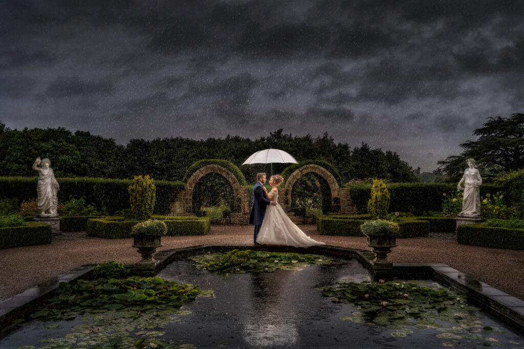 Couple embracing under umbrella in rain in the memorial garden at Allerton castle