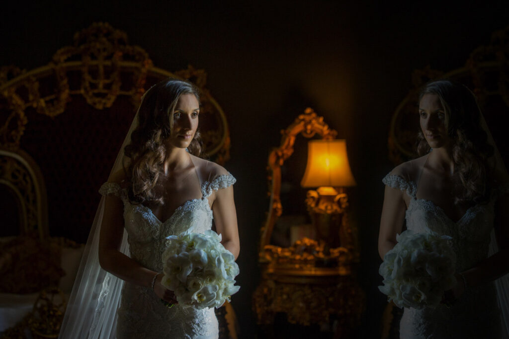 Bride with bouquet near mirror in dimly lit room.