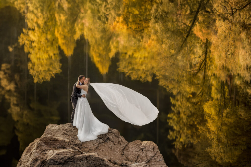 Bride and groom embracing on rock by reflective lake in the lake district