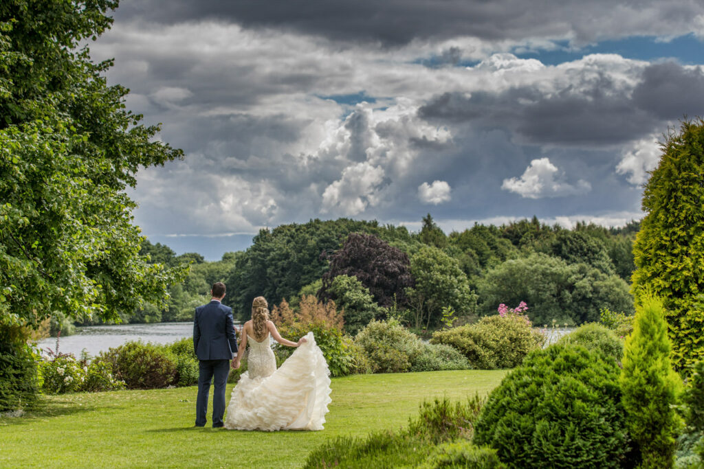 Bride and groom walking in garden by lake.