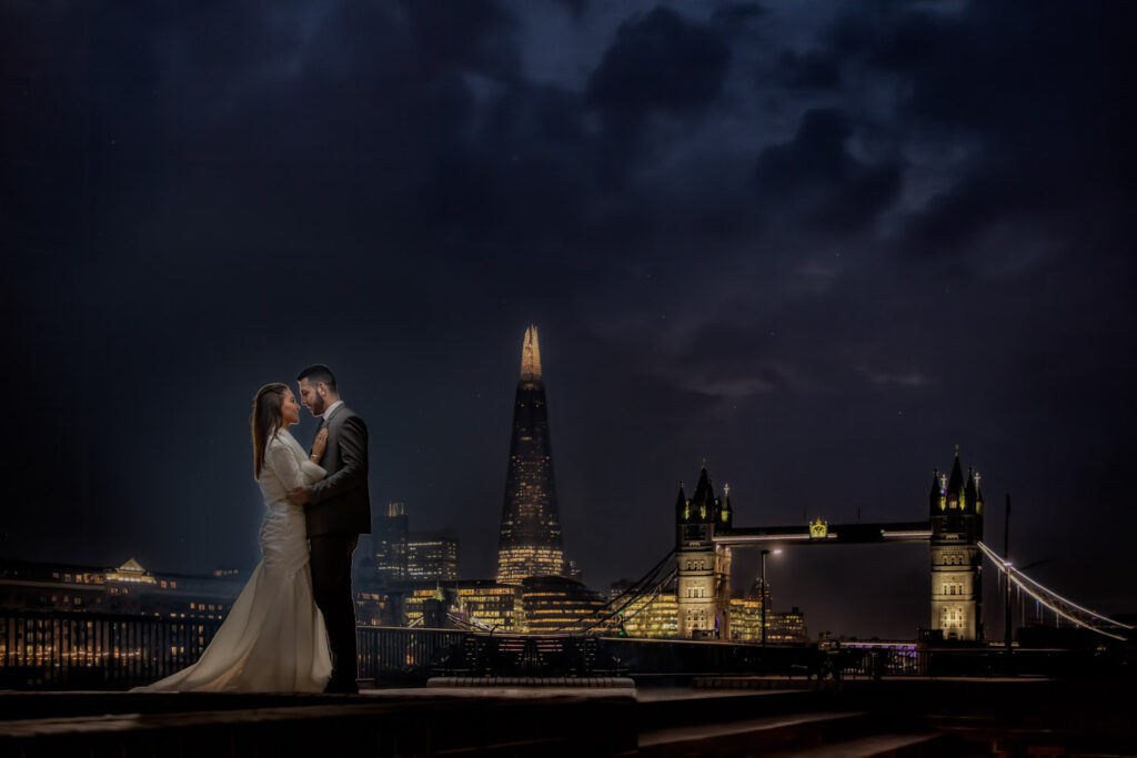 Bride and groom embracing, city skyline at night with illuminated bridge.