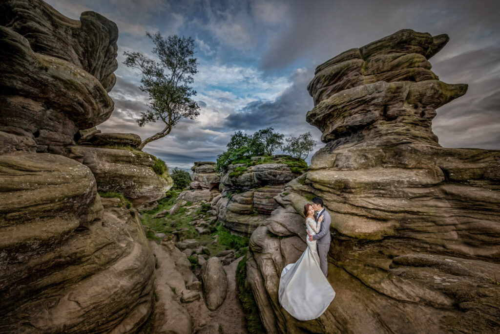 Couple embraces among dramatic rock formations under cloudy sky. Chris Chambers Yorkshire wedding photographer
