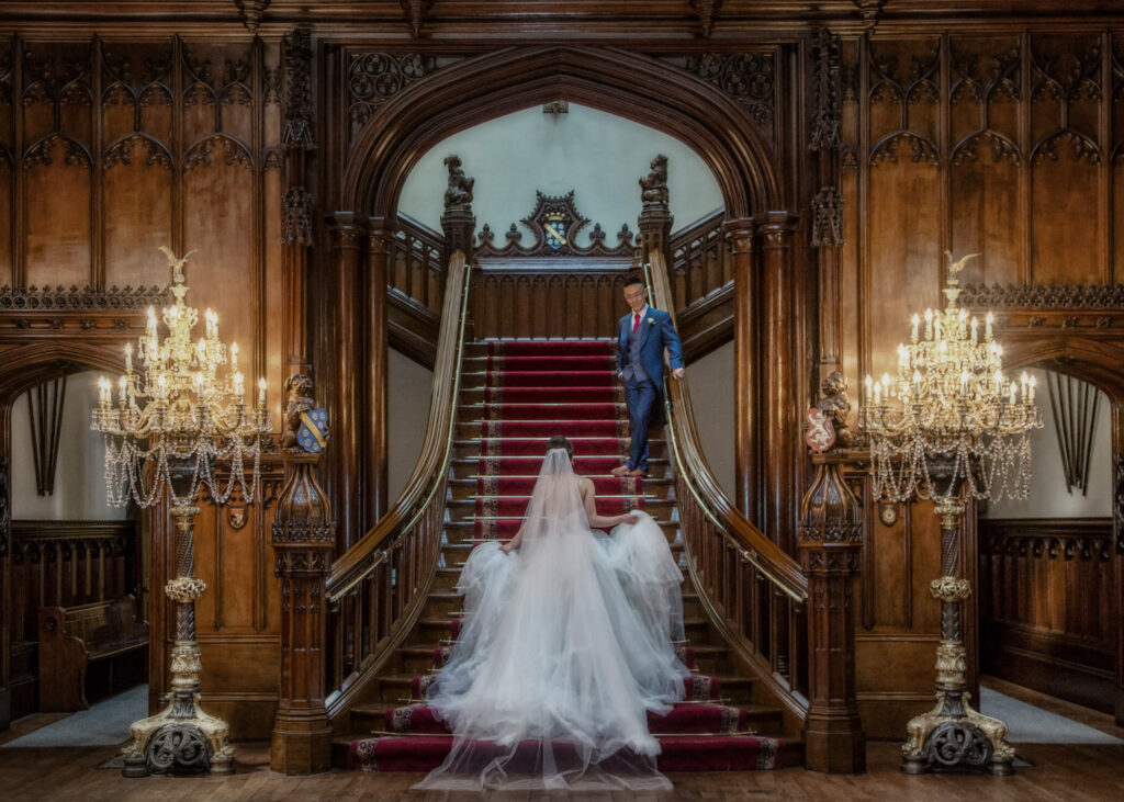 Bride ascending grand staircase with groom waiting at Allerton Castle.