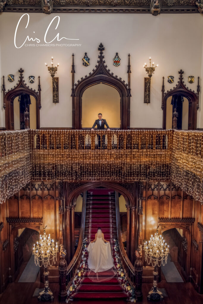 Bride ascending staircase, groom awaits in ornate hall.