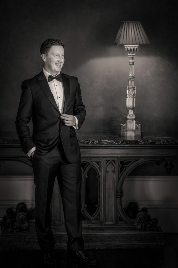Man in black tuxedo smiling beside vintage lamp. Allerton Castle wedding photographs