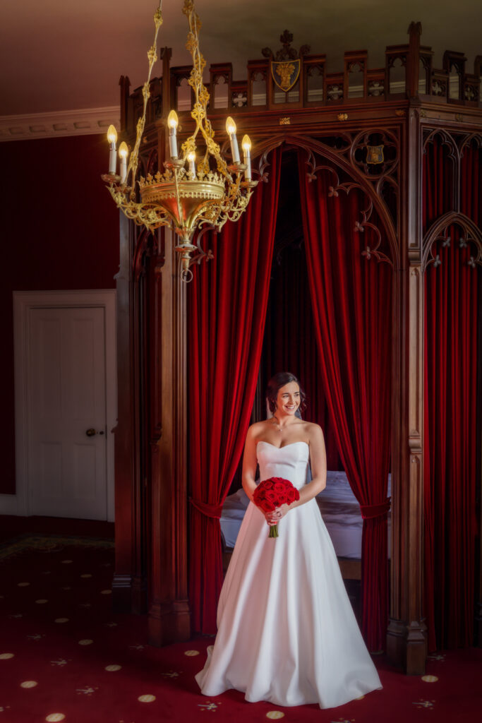 Bride in white dress with bouquet, elegant chandelier, red drapes.